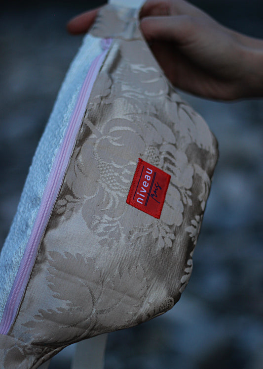 Banane Tchikita en damas beige, sangle blanche, zip rose, logo sur le devant, zoom sur le logo.