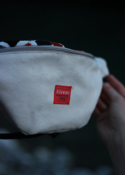 Banane Tchikita en suédine beige, sangle noire, zip gris, logo sur le devant, zoom sur le logo.