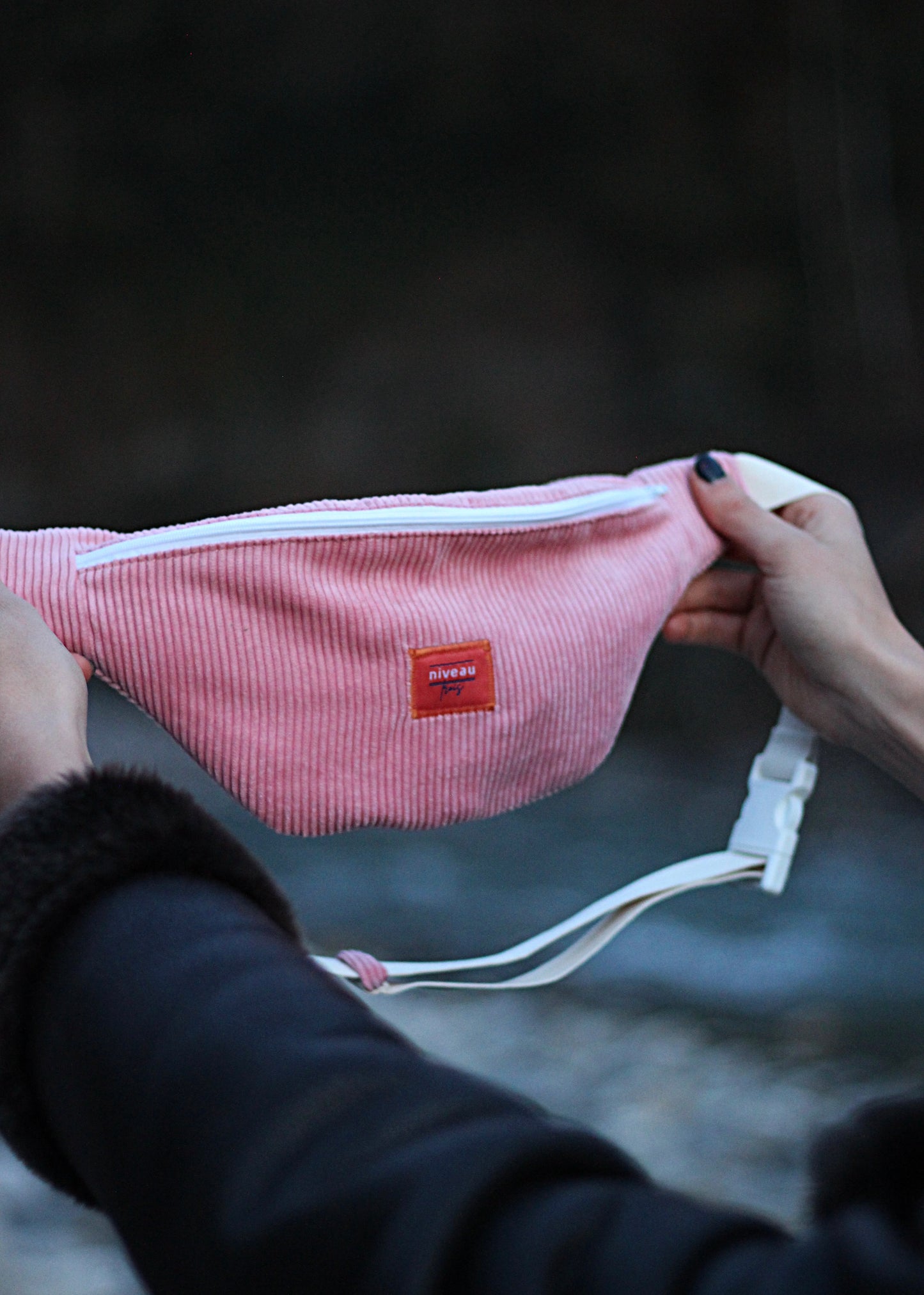 Banane Tchikita en velour côtelé rose, sangle blanche, zip blanc, logo sur le devant, vue bras tendu.